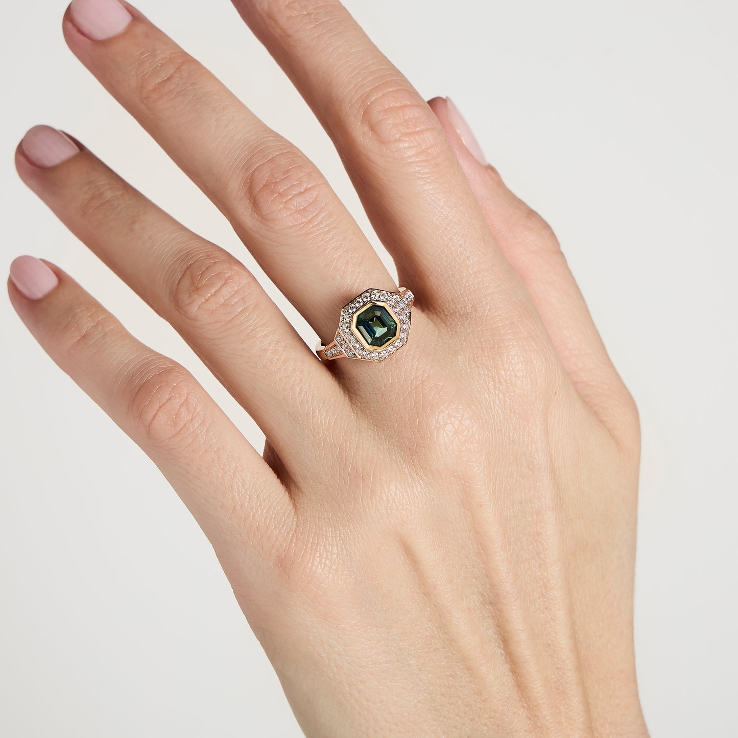 Une main à la peau claire porte la superbe Bague Saphir Teal • 1,56 ct de Bonnot Paris, ornée d'un saphir bleu-vert hexagonal. La pierre précieuse est entourée de petits diamants blancs, élégamment sertis sur un anneau en or. Les ongles sont manucurés en rose pâle.
