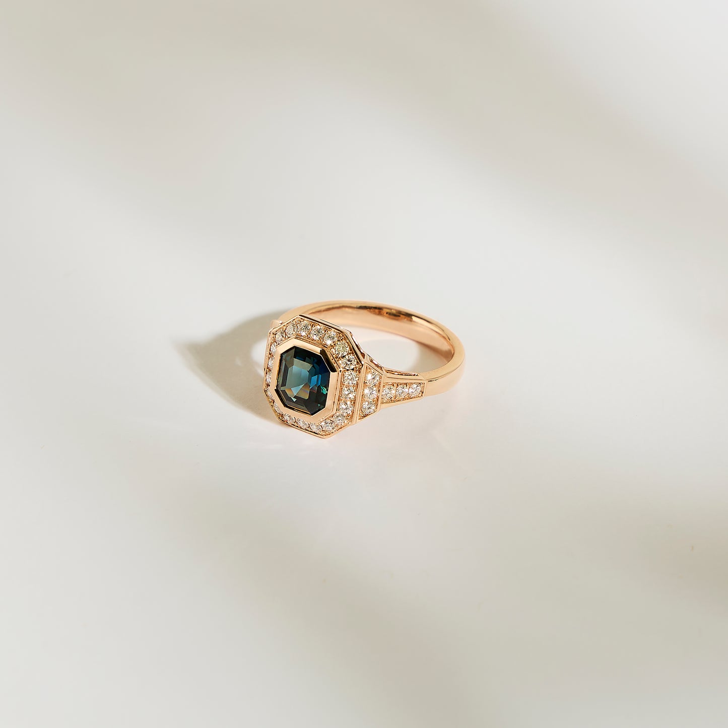 La Bague Saphir Teal de 1,56 carat signée Bonnot Paris arbore un saphir bleu-vert octogonal central entouré de petits diamants blancs. Elle repose élégamment sur une surface lisse et pâle, dont les ombres douces accentuent le design.