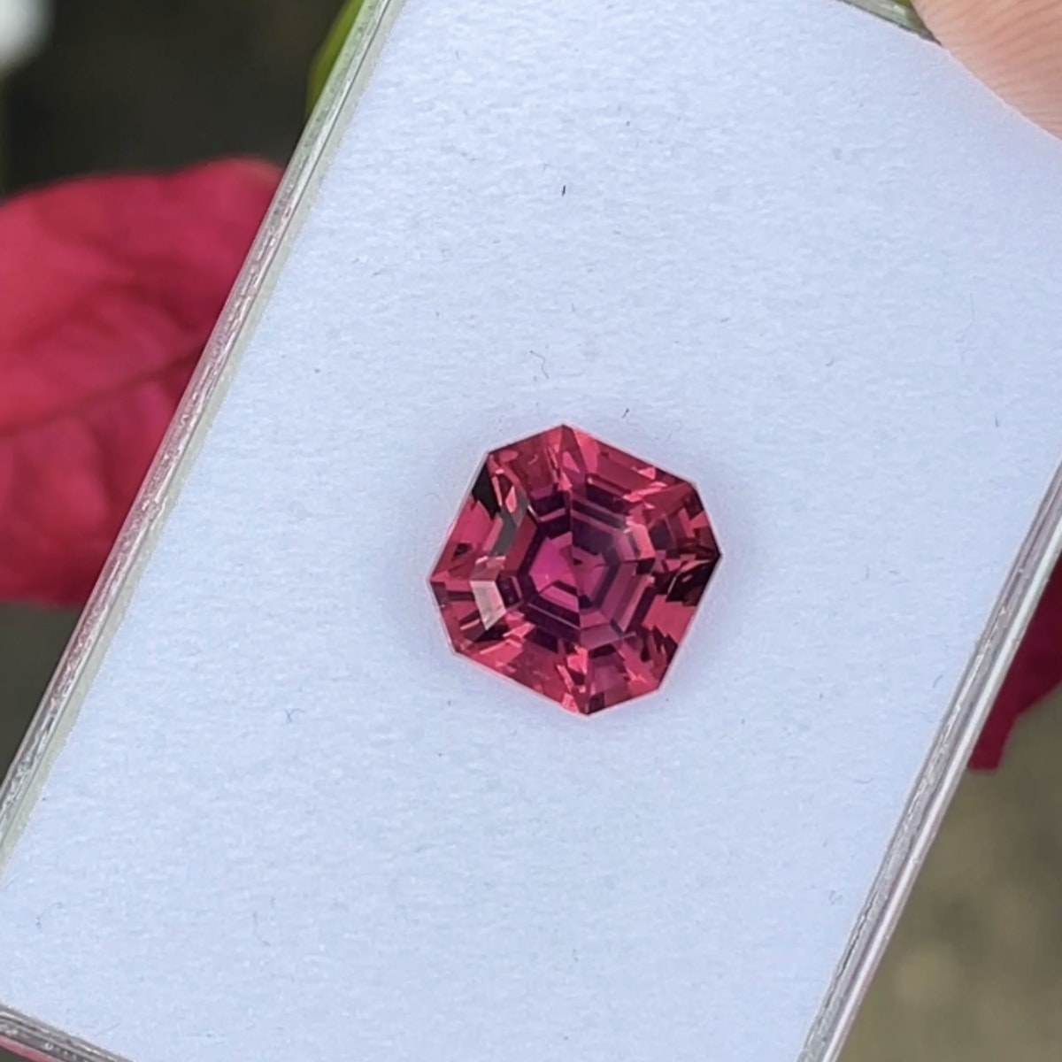 Une tourmaline rose hexagonale de 6,79 carats signée Bonnot Paris repose sur un fond blanc, ses facettes d'un rose éclatant étincelant. Une main la tient délicatement par le bord, avec un objet rouge flou en arrière-plan.