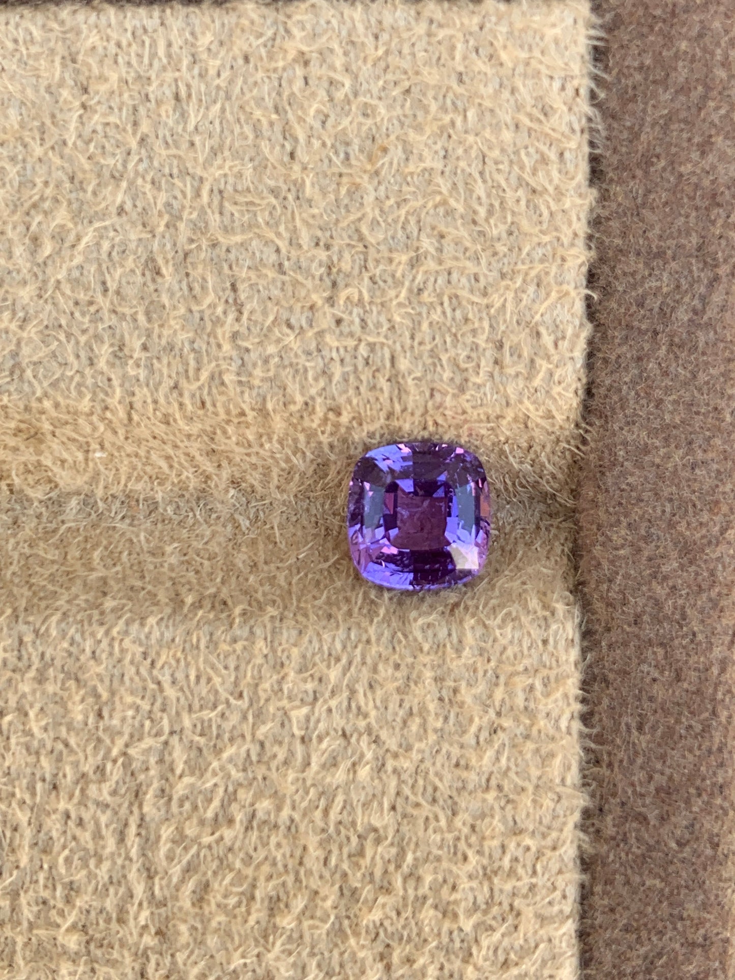 Le Saphir Violet • 1,5ct de Bonnot Paris, une superbe pierre précieuse violette vibrante de Madagascar, repose sur un tissu beige texturé, mettant en valeur ses facettes scintillantes et sa couleur riche rappelant une pierre lumineuse.