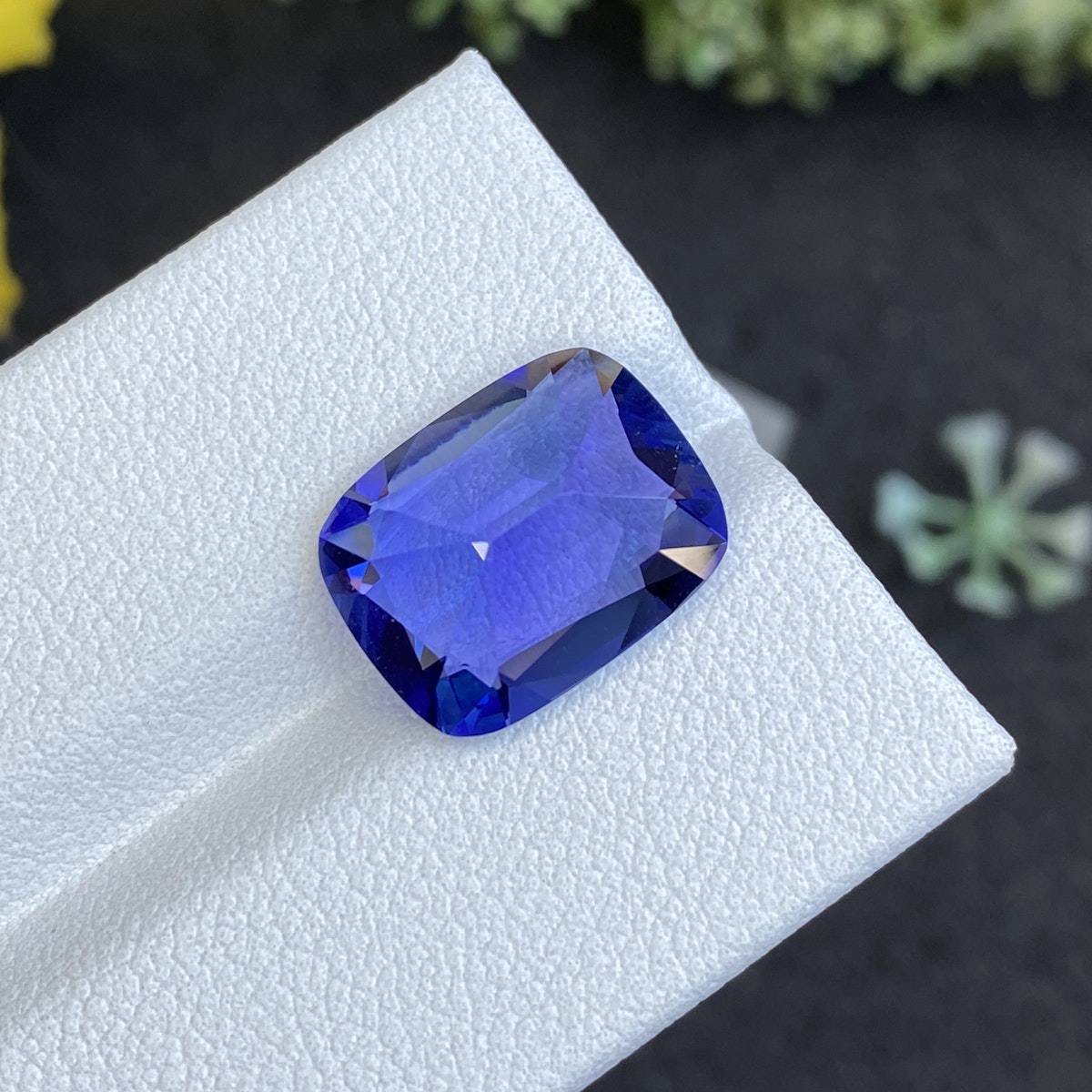 Un gros plan de la Tanzanite taille coussin de 6,66 ct de Bonnot Paris montre sa teinte bleue vibrante et ses facettes réfléchissantes, sur fond de surface texturée blanche avec des éléments verts flous en arrière-plan.