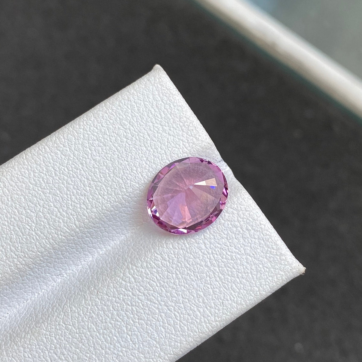 Présenté sur une surface blanche texturée sur fond noir, le Spinelle Rose · 3,93 ct de Bonnot Paris présente une taille ovale avec un bord brillant à proximité.