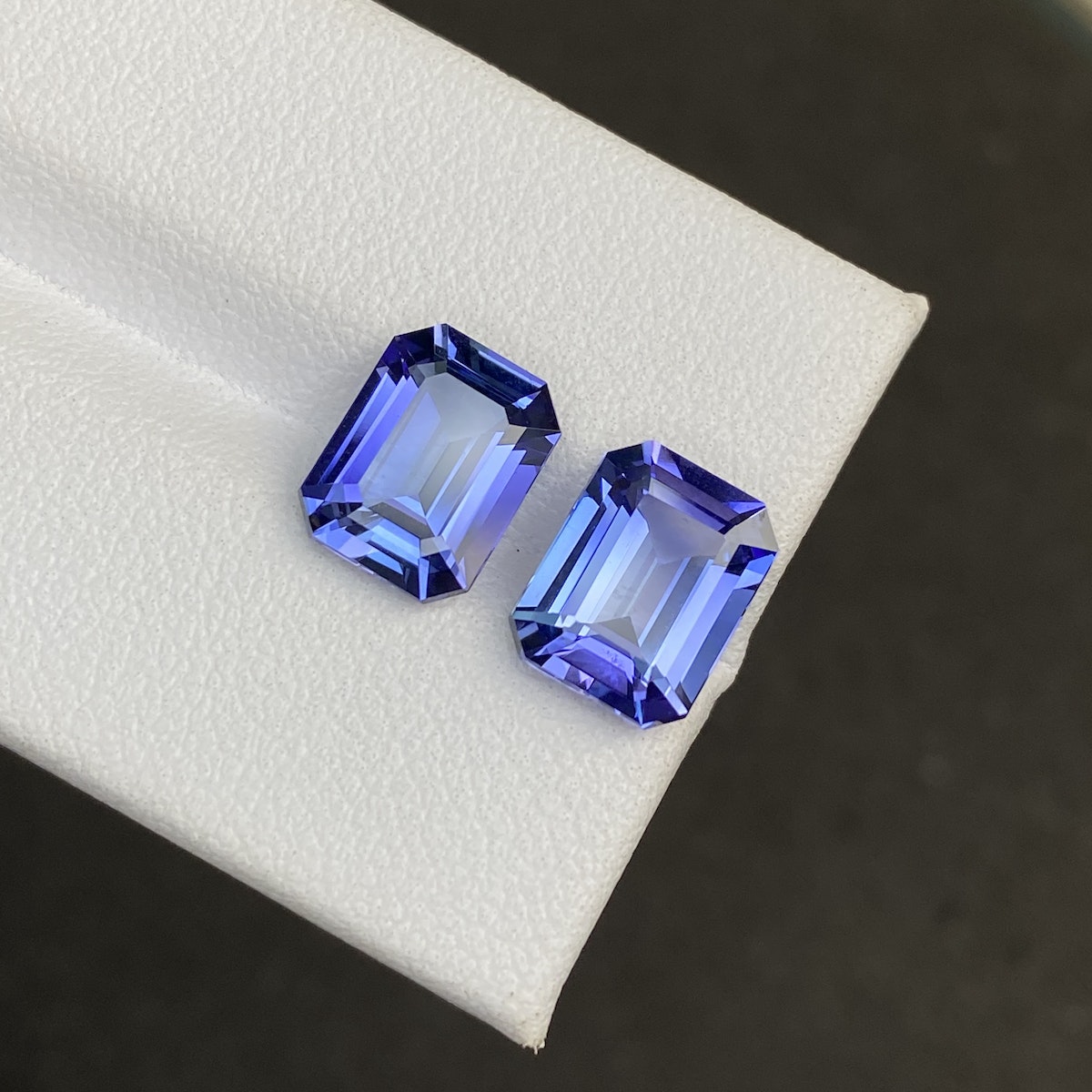 Deux tanzanites Bonnot Paris taille émeraude (5,82 ct chacune) sont placées côte à côte sur une surface blanche, présentant des couleurs vibrantes et des facettes précises sur un fond noir contrastant.