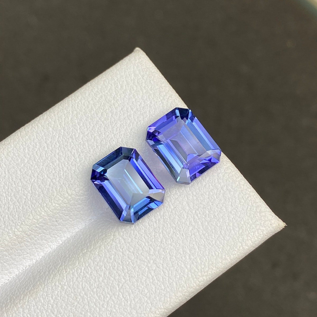 Deux tanzanites Bonnot Paris, de 5,82 carats chacune, de taille émeraude rectangulaire et aux reflets bleu-violet, sont posées sur une surface blanche texturée. Elles présentent un dégradé de couleurs saisissant sous la lumière réfléchie sur un fond neutre et flou.