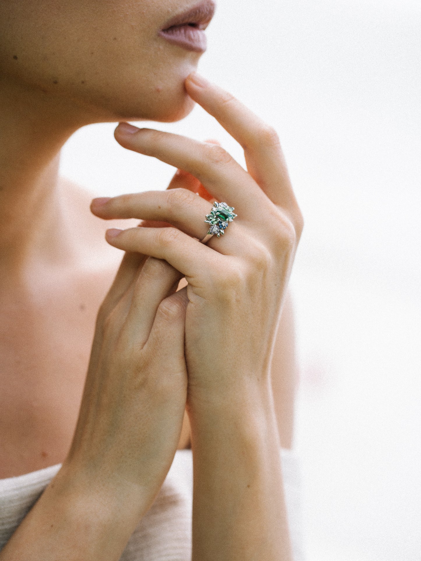 Un gros plan montre une personne touchant son menton avec des doigts élégants ornés de la bague scintillante « Green Bloom - Tsavorite » de Bonnot Paris comportant une grande pierre précieuse Grenat tsavorite, sertie sur un fond légèrement flou qui met en valeur ce bijou exquis.