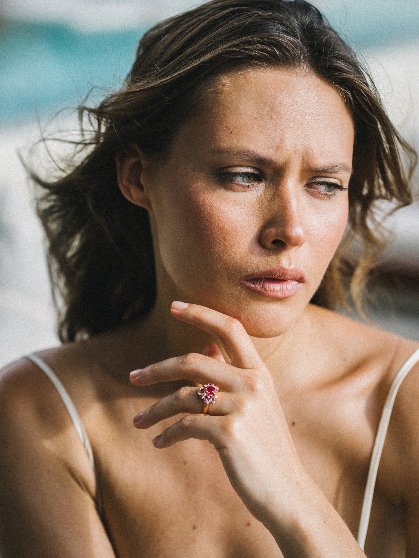 Une femme aux cheveux ondulés regarde pensivement de côté, le menton posé sur sa main ornée de la bague Bonnot Paris « Rose Bloom » sertie d'un spinelle Mahenge éclatant. Elle porte un haut à fines bretelles, encadré par un arrière-plan légèrement flou qui accentue son humeur contemplative.