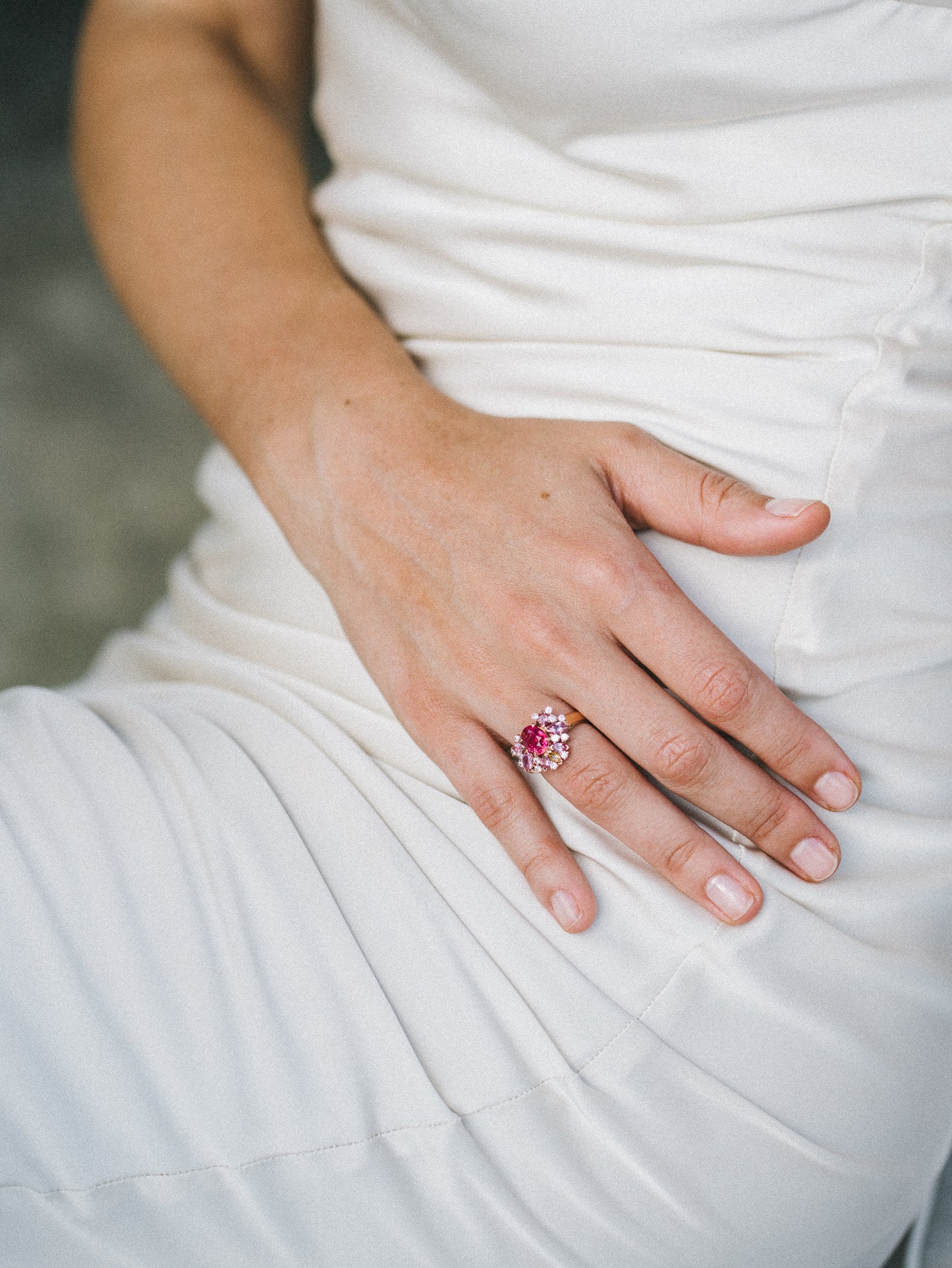 Une personne vêtue d'une robe blanche pose subtilement sa main sur ses genoux, mettant en valeur la bague Rose Bloom - Spinelle Mahenge de Bonnot Paris. Sertie d'une magnifique pierre rose entourée de petits diamants, l'arrière-plan délicatement flou met en valeur les détails raffinés de la bague.