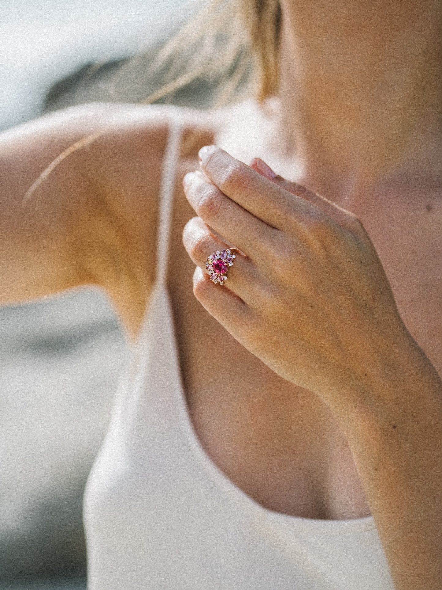 Une femme vêtue d'un haut blanc sans manches modèle sa main ornée du "Rose Bloom - Spinelle Mahenge" de Bonnot Paris, positionnée près de son épaule tandis que la lumière du soleil projette une lueur chaleureuse sur un arrière-plan extérieur flou.