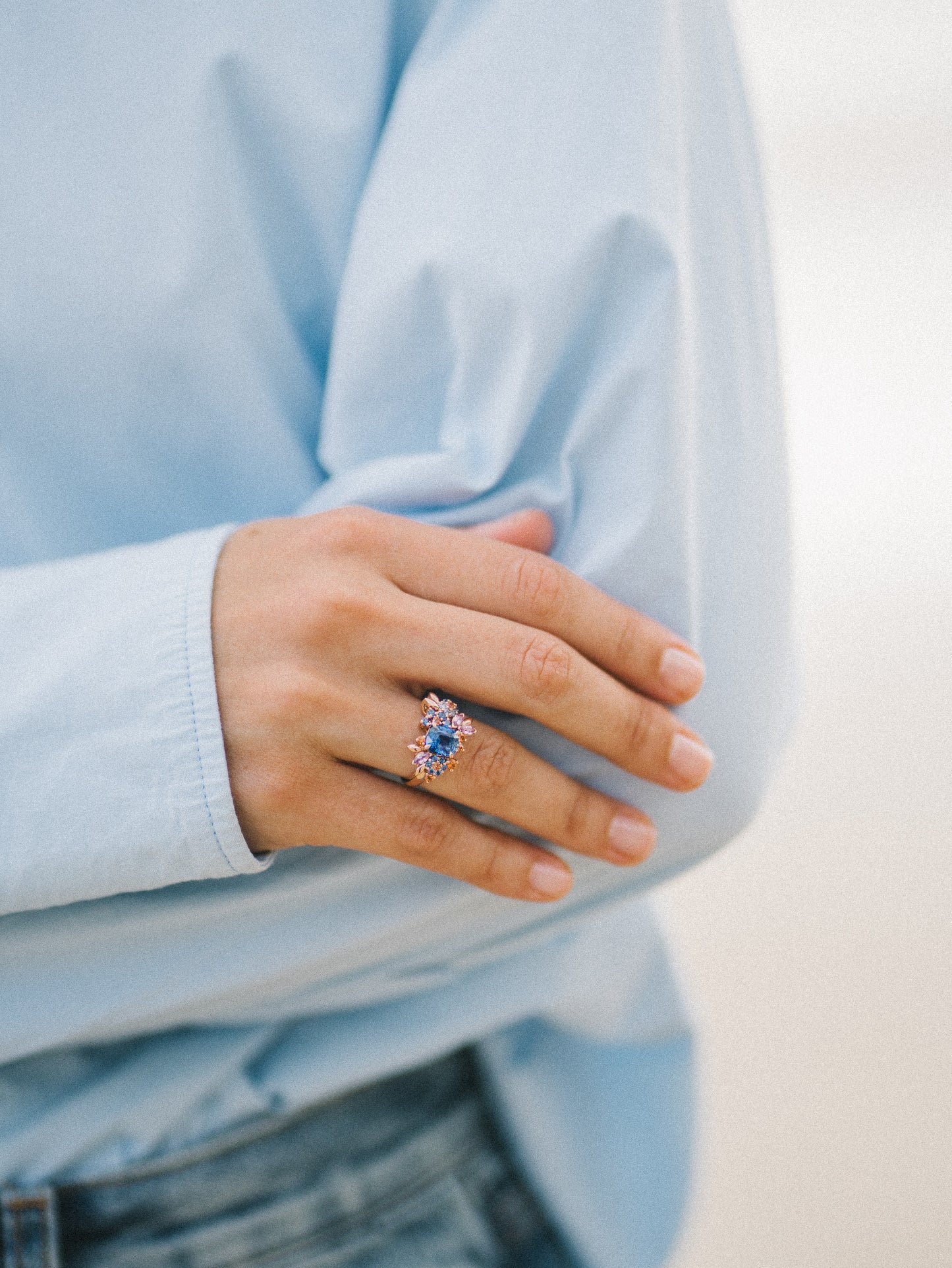 Une personne en chemise bleue et jean met en valeur sa main avec la bague Navy Bloom - Saphir de Bonnot Paris, comportant un superbe saphir bleu entouré de plus petites pierres précieuses, tandis que son bras se croise gracieusement sur l'autre.