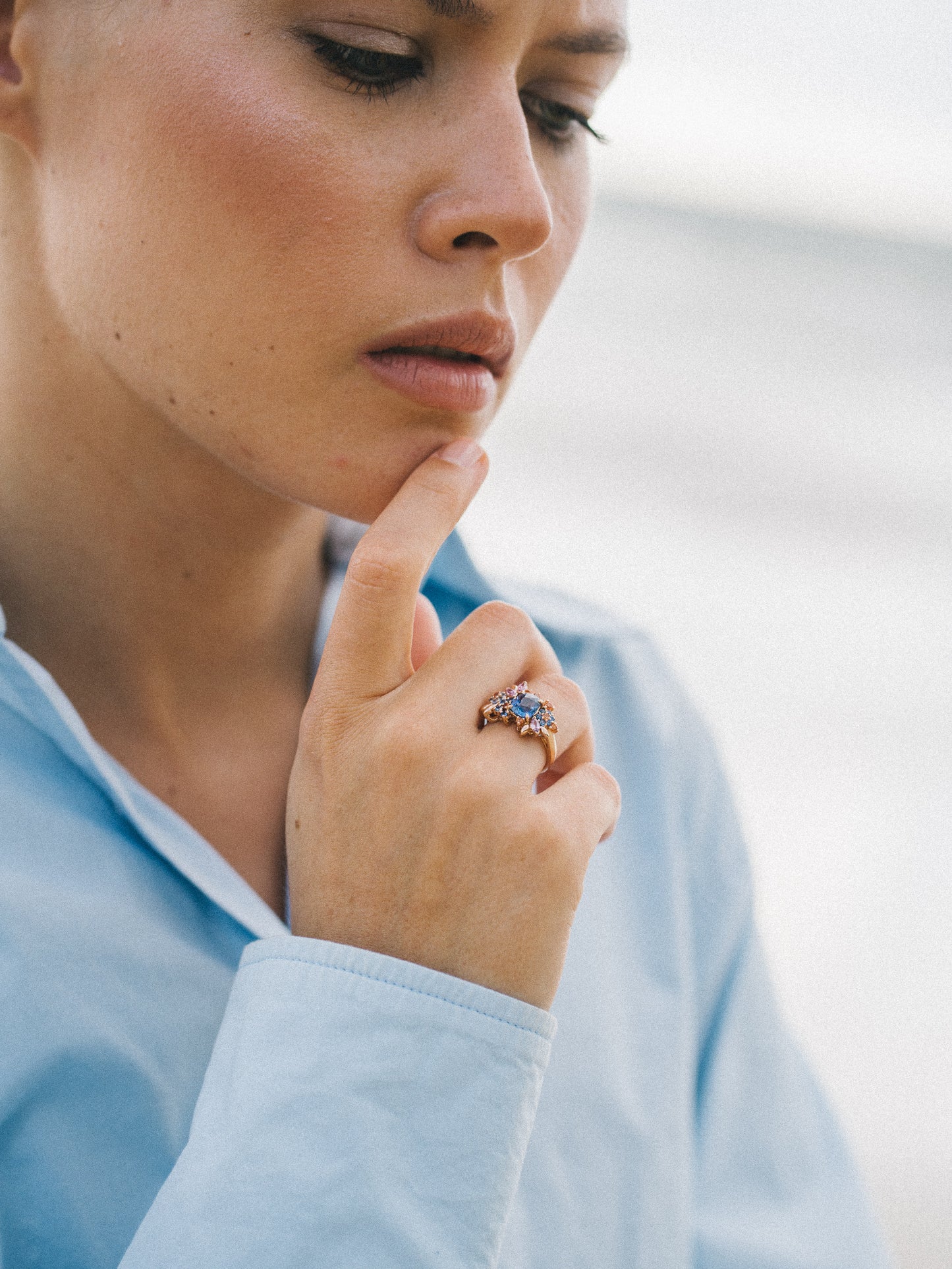 Une personne, vêtue d'une chemise bleu clair, se touche pensivement le menton. Son index gauche est orné de la bague Navy Bloom - Saphir de Bonnot Paris. L'arrière-plan évoque un décor extérieur.