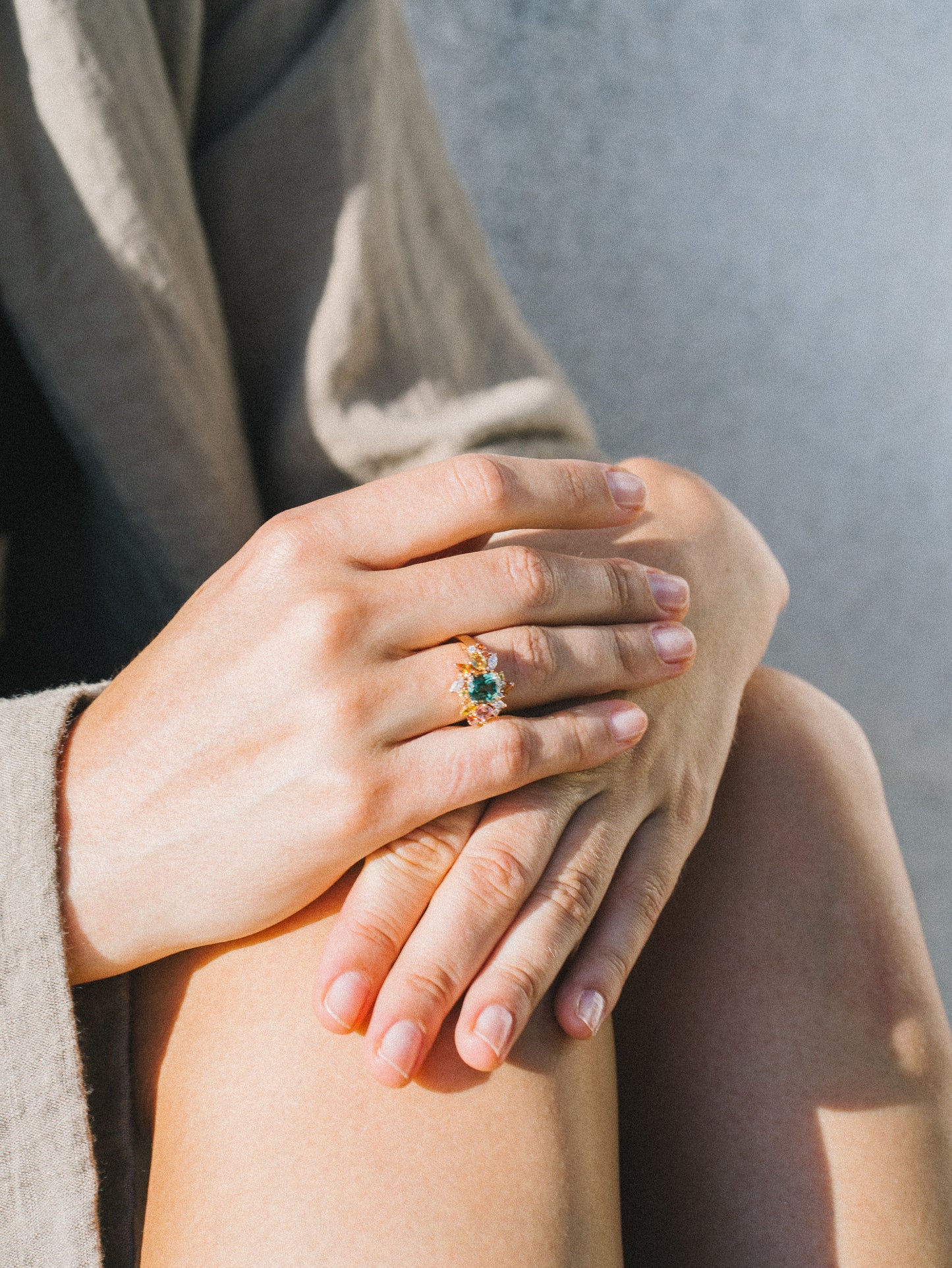 Une personne pose sa main gauche sur son genou, portant la bague Sea Bloom - Tourmaline de Bonnot Paris, ornée de délicats diamants. Sur un arrière-plan légèrement flou, la lumière du soleil projette une douce lueur sur son vêtement clair, soulignant l'élégance de la bague.