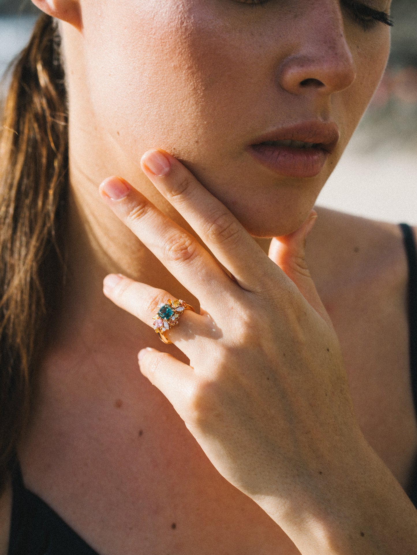 Une personne à la peau claire pose son menton sur sa main, mettant en valeur la bague Sea Bloom - Tourmaline de Bonnot Paris. Ses longs cheveux bruns sont attachés en arrière et elle porte un haut noir. L'image est doucement éclairée par la lumière naturelle, créant une atmosphère sereine.
