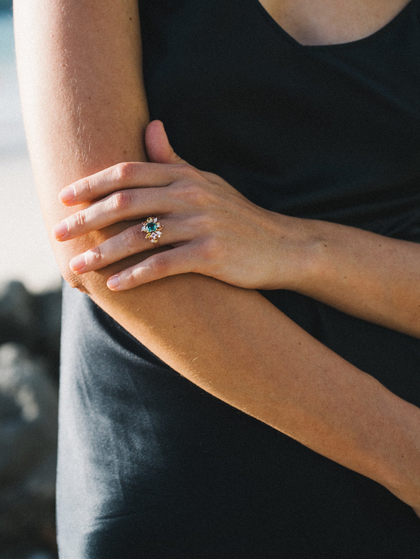 Une personne vêtue d'une robe noire sans manches se tient à l'extérieur, les bras croisés, présentant la bague Sea Bloom - Tourmaline de Bonnot Paris sur sa main droite ; l'accent est mis sur la superbe bague et les mains.