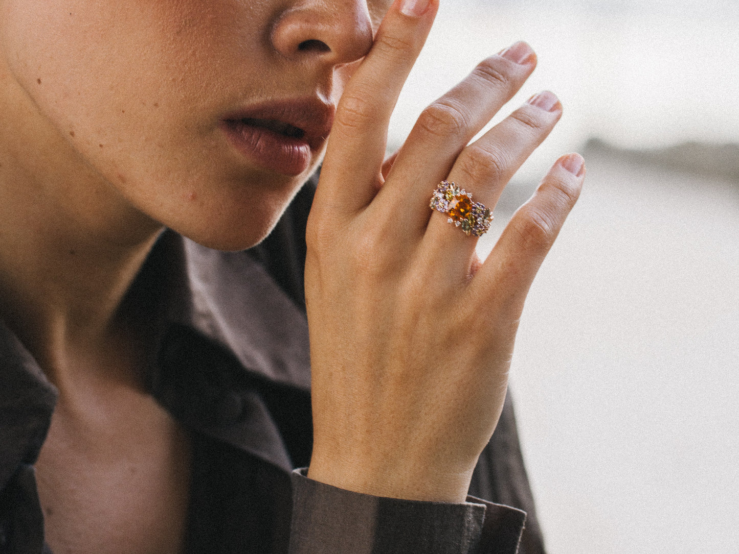 En gros plan, une personne en chemise marron touche légèrement son visage orné de la bague Golden Bloom - Grenat Mandarin de Bonnot Paris, son éblouissant grenat spessartite et diverses pierres précieuses créant une floraison de couleurs vives sur le fond flou.