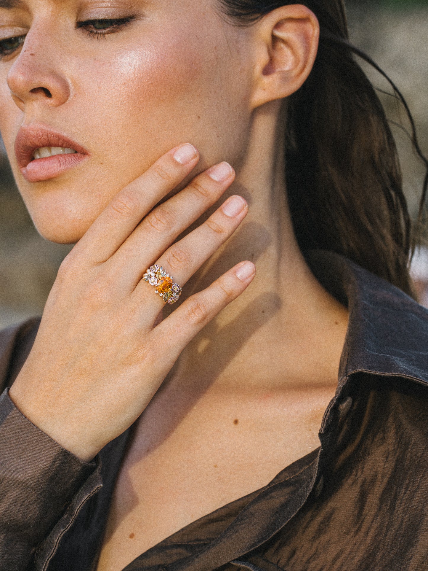 Une personne, cheveux mouillés et maquillage léger, se touche le visage, mettant en valeur une bague ornée de pierres précieuses qui rappelle l'élégance du Golden Bloom – Grenat Mandarin de Bonnot Paris. Elle porte un chemisier sombre transparent sur un fond légèrement flou, suggérant un décor extérieur subtilement sophistiqué.