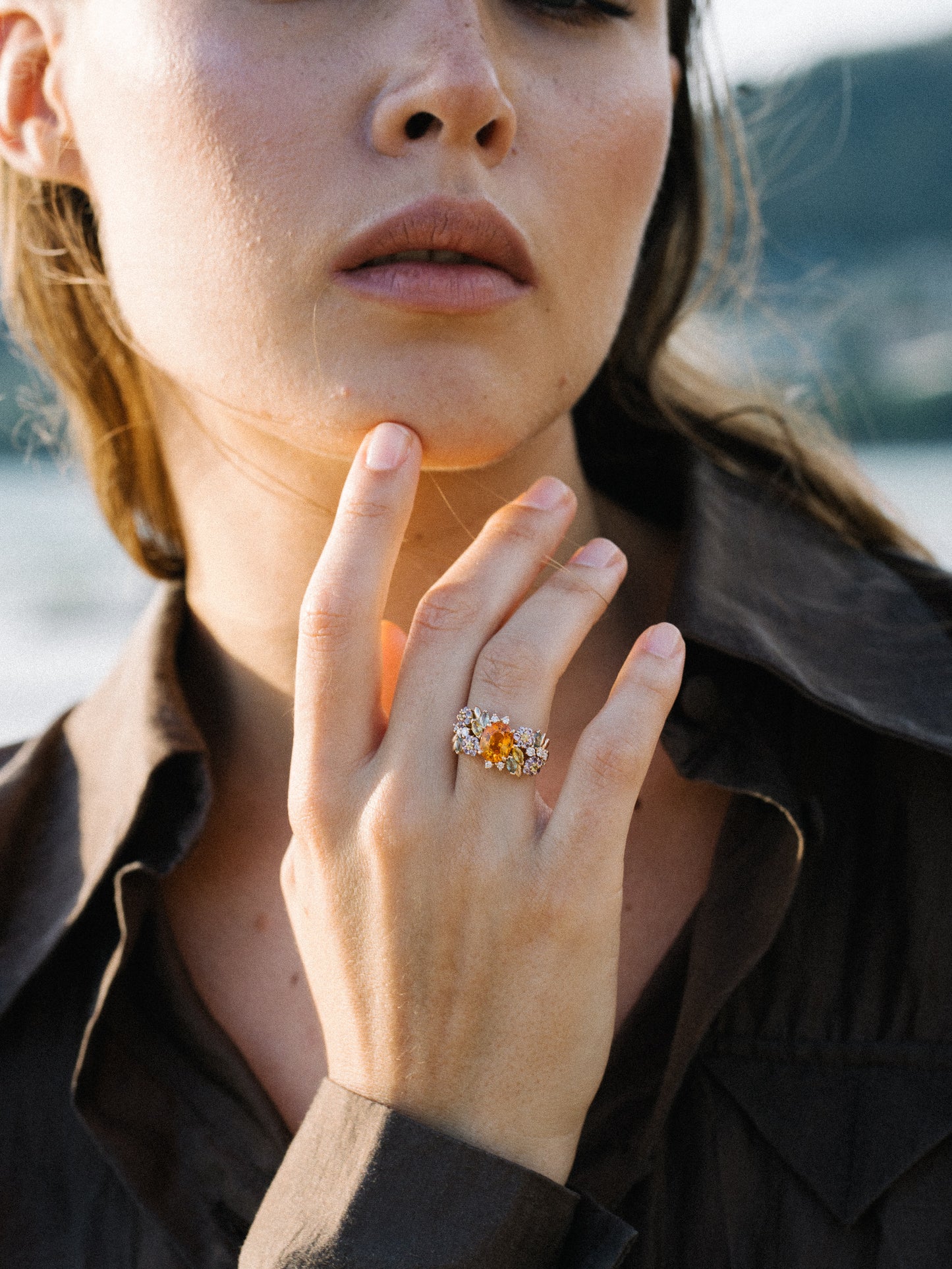 Une personne en chemise sombre dévoile sa main, élégamment ornée de la bague Golden Bloom - Grenat Mandarin de Bonnot Paris, ornée d'une pierre centrale ambrée entourée de plus petites pierres transparentes. Le doux flou de l'arrière-plan extérieur évoque les tons de Color Blossom.