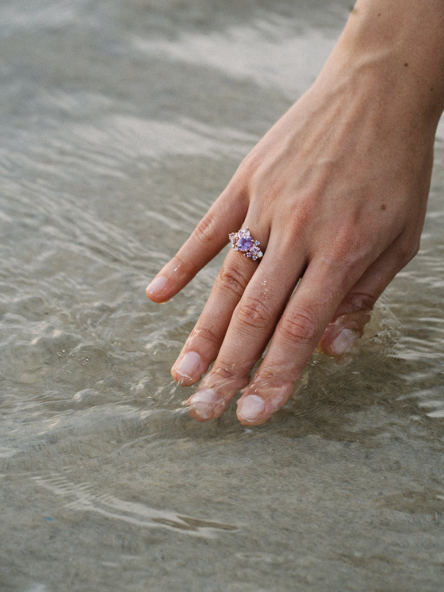 Une main portant la bague Purple Bloom - Saphir de Bonnot Paris effleure légèrement l'eau claire, formant de douces ondulations.