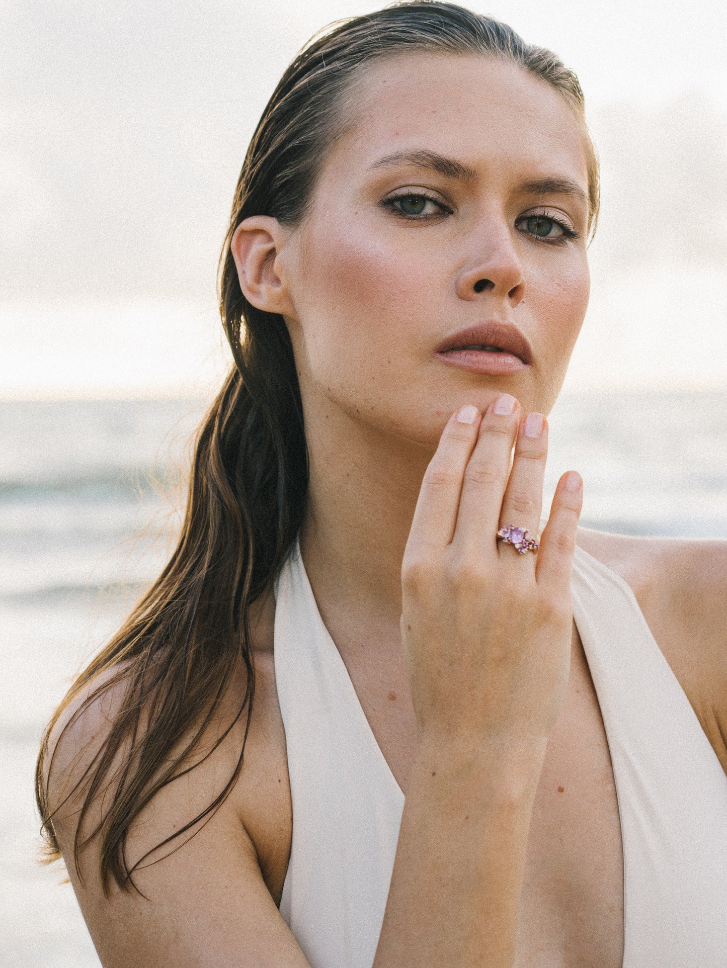 Une femme aux cheveux mouillés pose pensivement, sa main près de son visage affichant la bague Purple Bloom – Saphir de Bonnot Paris. Elle porte un haut blanc dos nu sur fond flou de vagues océaniques sereines.