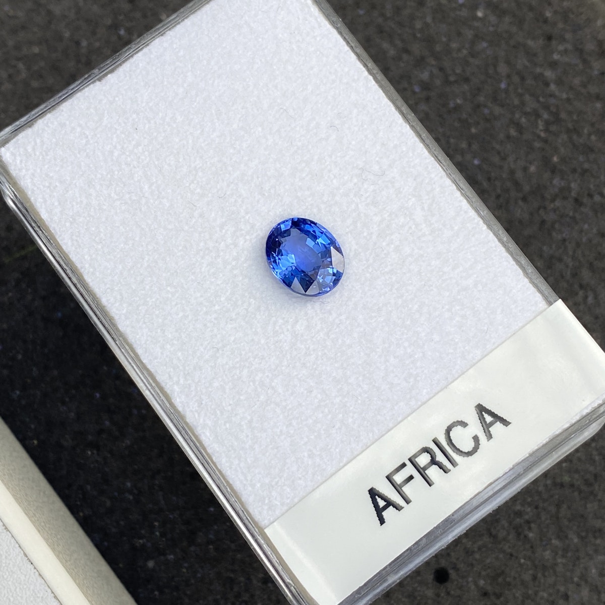 Une petite pierre précieuse bleue ovale est présentée sur un support blanc dans un écrin transparent étiqueté « Saphir Bleu · 2,04 ct » par Bonnot Paris. Le fond sombre met en valeur la brillance de la pierre et de son écrin.