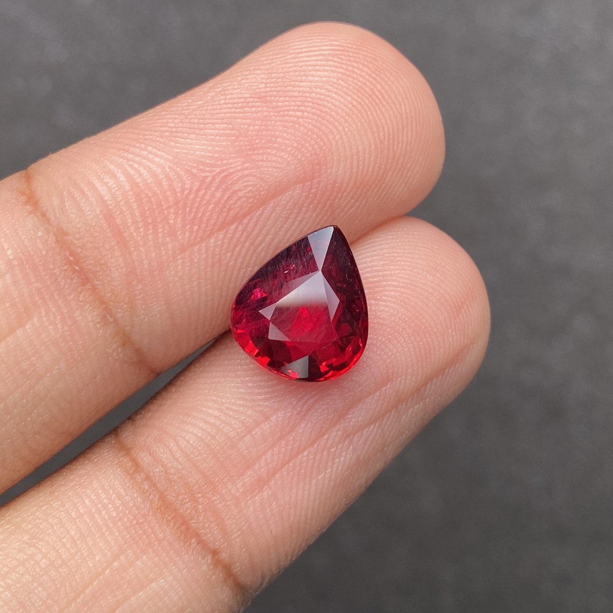 Une main tient le Rubis du Mozambique · 4,03 ct de Bonnot Paris, une pierre précieuse rouge foncé en forme de poire, entre le pouce et l'index sur un fond sombre et flou.