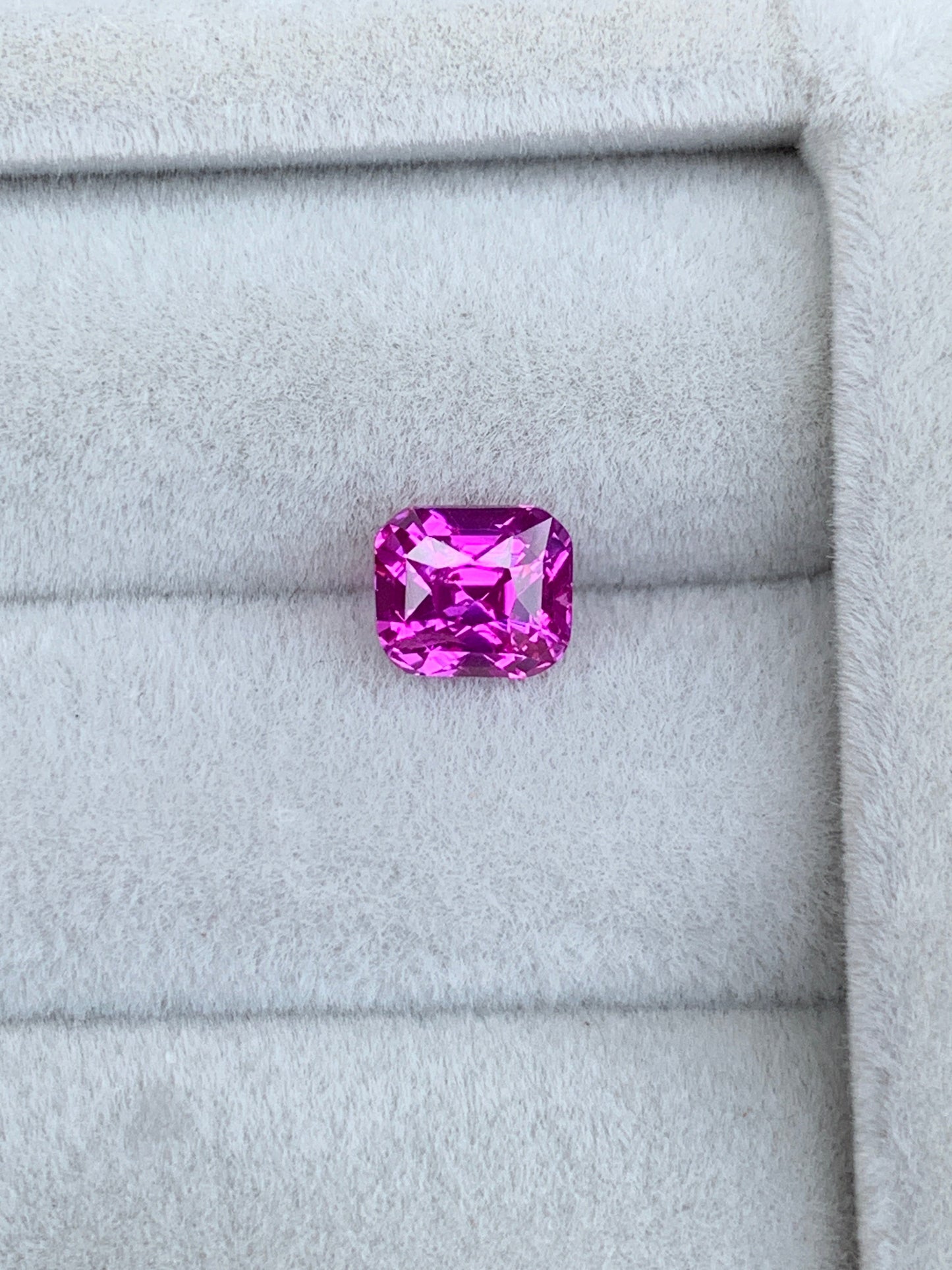Un Saphir Rose éclatant de 1,6 carat signé Bonnot Paris repose sur un tissu gris doux, avec une taille rectangulaire aux bords biseautés. Ses facettes reflètent brillamment la lumière, sublimant sa couleur vive : un véritable bijou sur mesure.