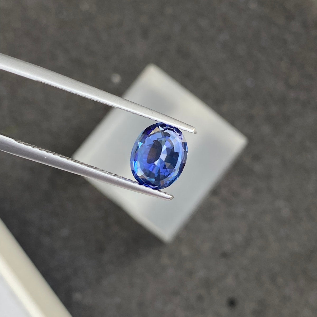 Une pince à épiler tient le Saphir Bleu · 2,04 ct, une pierre précieuse bleue taille ovale de Bonnot Paris, sur un fond gris flou. La pierre facettée présente différentes nuances de bleu.