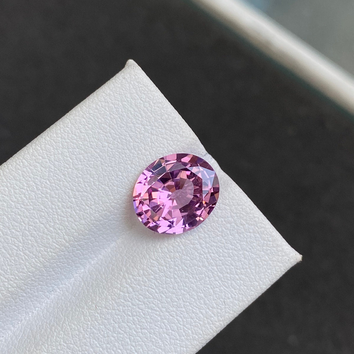 Un spinelle rose ovale facetté de 3,93 ct de Bonnot Paris est présenté sur un fond texturé blanc, avec une teinte rose vibrante et des coupes complexes qui reflètent la lumière.