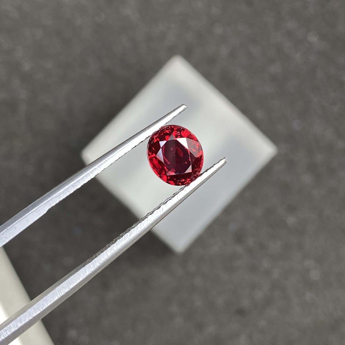 Une pince métallique maintient une pierre précieuse ronde facettée Spinelle Rouge · 1,25 carat de Bonnot Paris sur une surface texturée gris foncé. La pierre scintille de reflets lumineux, et un carré blanc flou apparaît en arrière-plan.