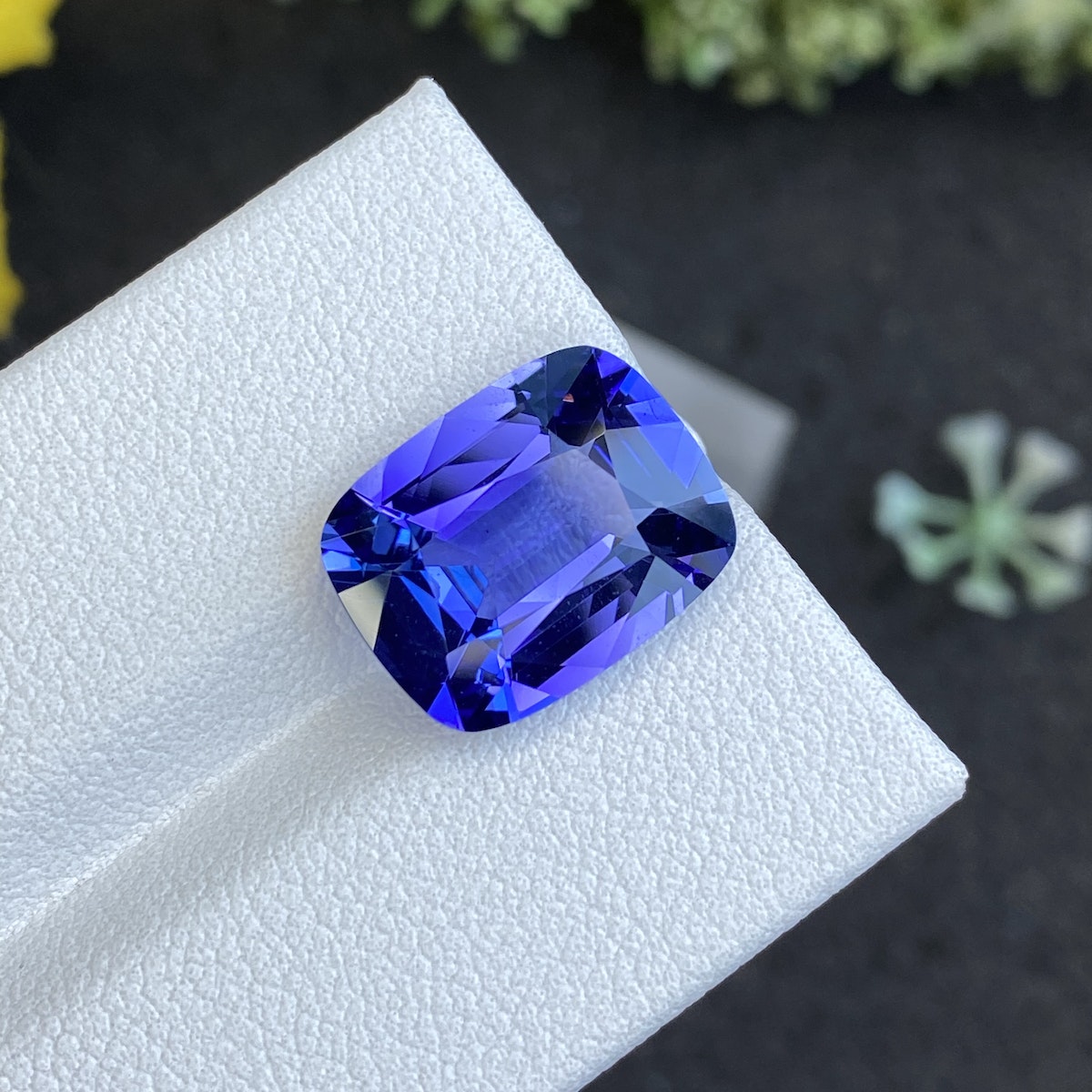 Une tanzanite facettée de 6,66 ct de Bonnot Paris brille de teintes bleues vibrantes sur une surface blanche, sur fond sombre avec des touches de verdure.