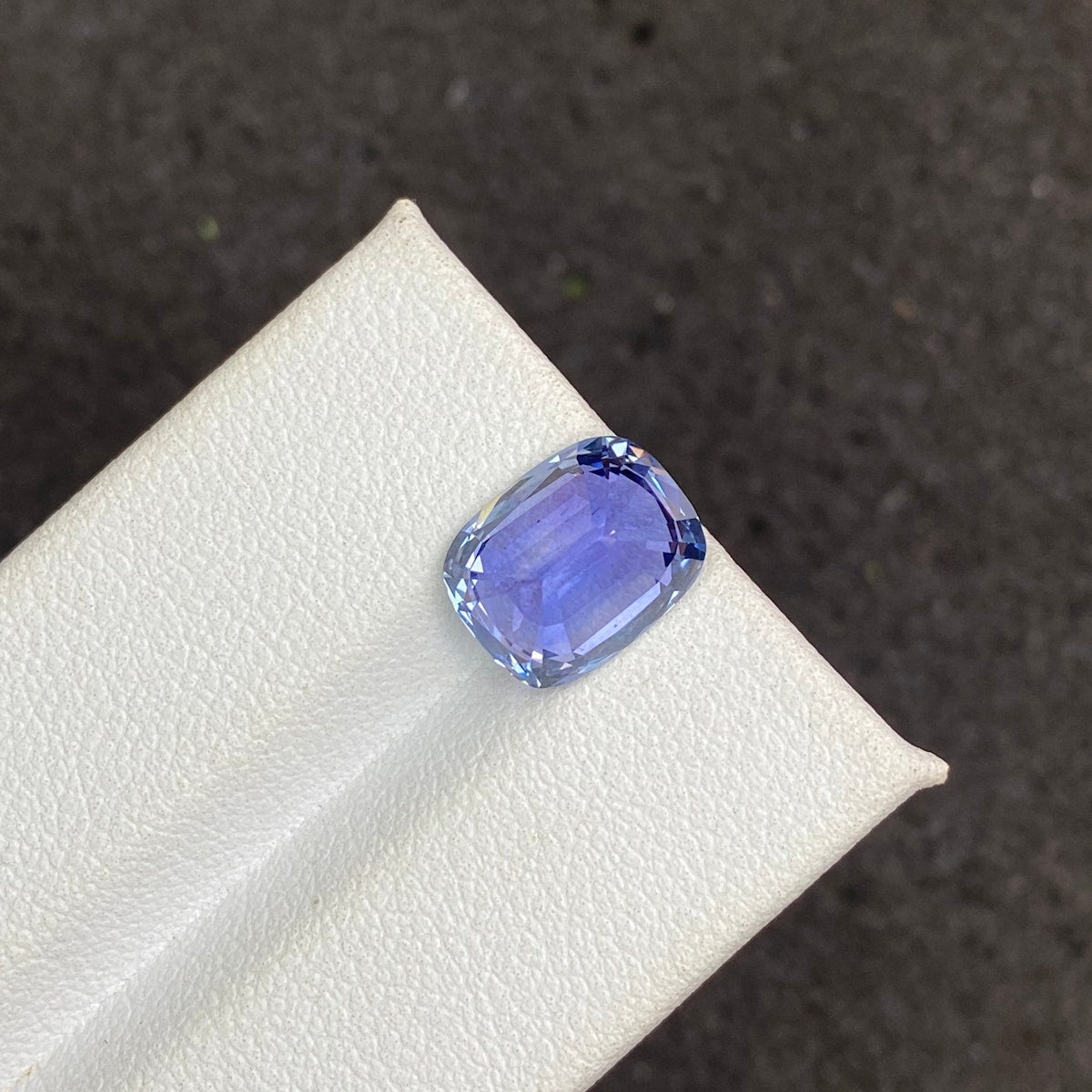 Gros plan sur le Saphir Bleu · 5,19 carats de Bonnot Paris, une pierre précieuse bleue taille coussin aux facettes brillantes sur une surface texturée beige clair. Le fond sombre et flou met en valeur sa couleur vive et son éclat.