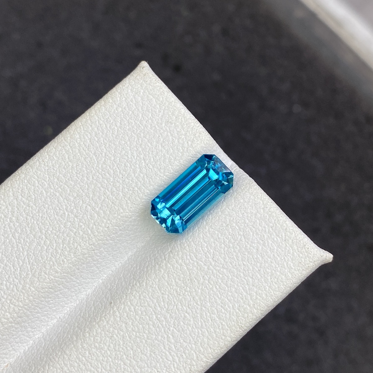 Le Zircon Bleu Bonnot Paris de 4,44 ct, avec sa taille rectangulaire éclatante, est mis en valeur sur une surface blanche texturée. Ses facettes bleues reflètent magnifiquement la lumière sur le fond sombre et flou, soulignant ainsi l'éclat de la pierre.