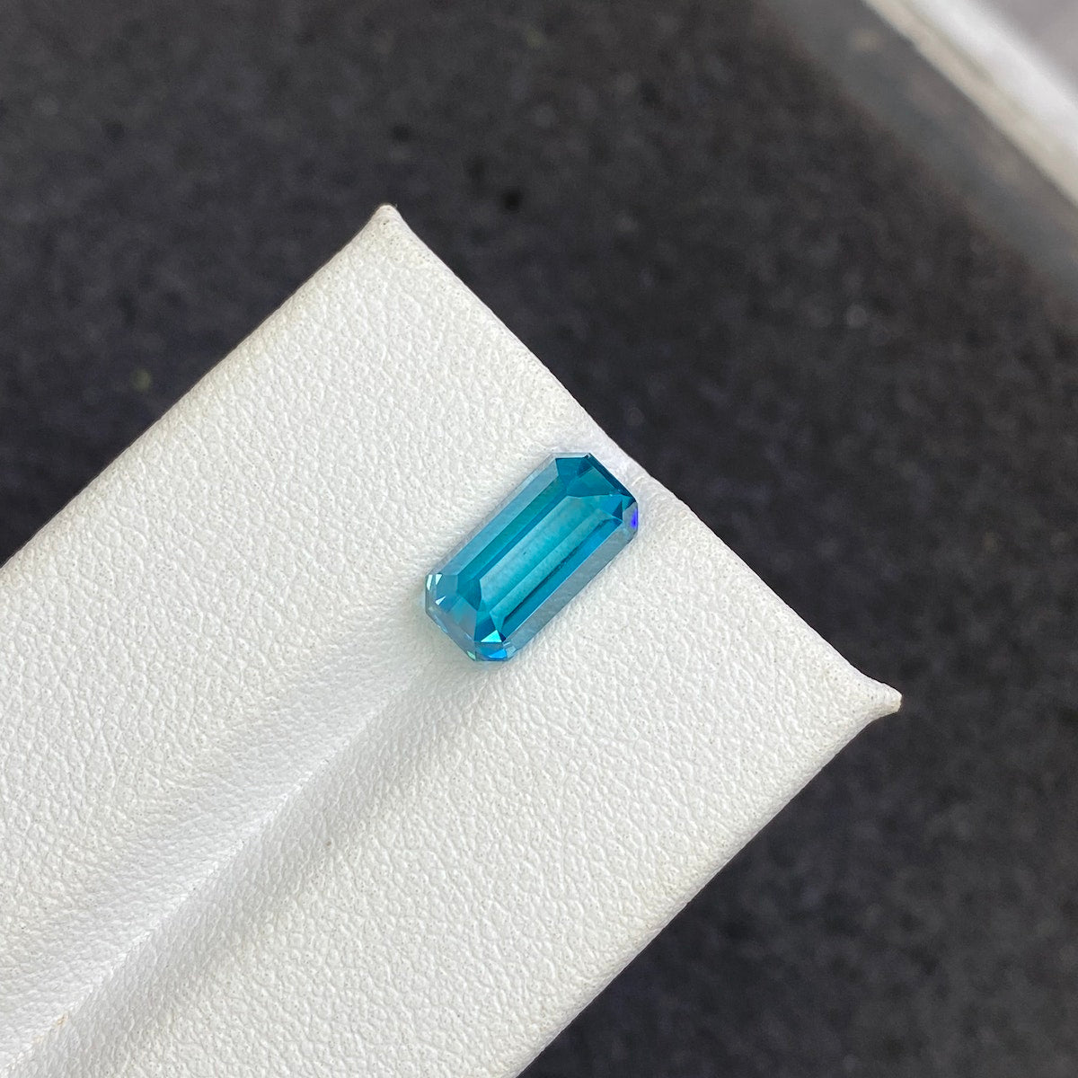 Une pierre précieuse rectangulaire taille émeraude Zircon Bleu · 4,44 ct de Bonnot Paris est présentée sur une surface texturée blanche sur un fond sombre.