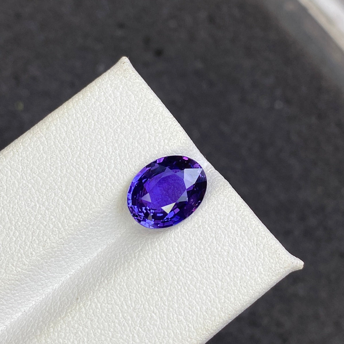Le Saphir Violet · 3,5ct de Bonnot Paris est une pierre précieuse de taille ovale, d'un bleu profond, aux facettes réfléchissant la lumière, reposant sur une surface texturée blanche sur un fond sombre.