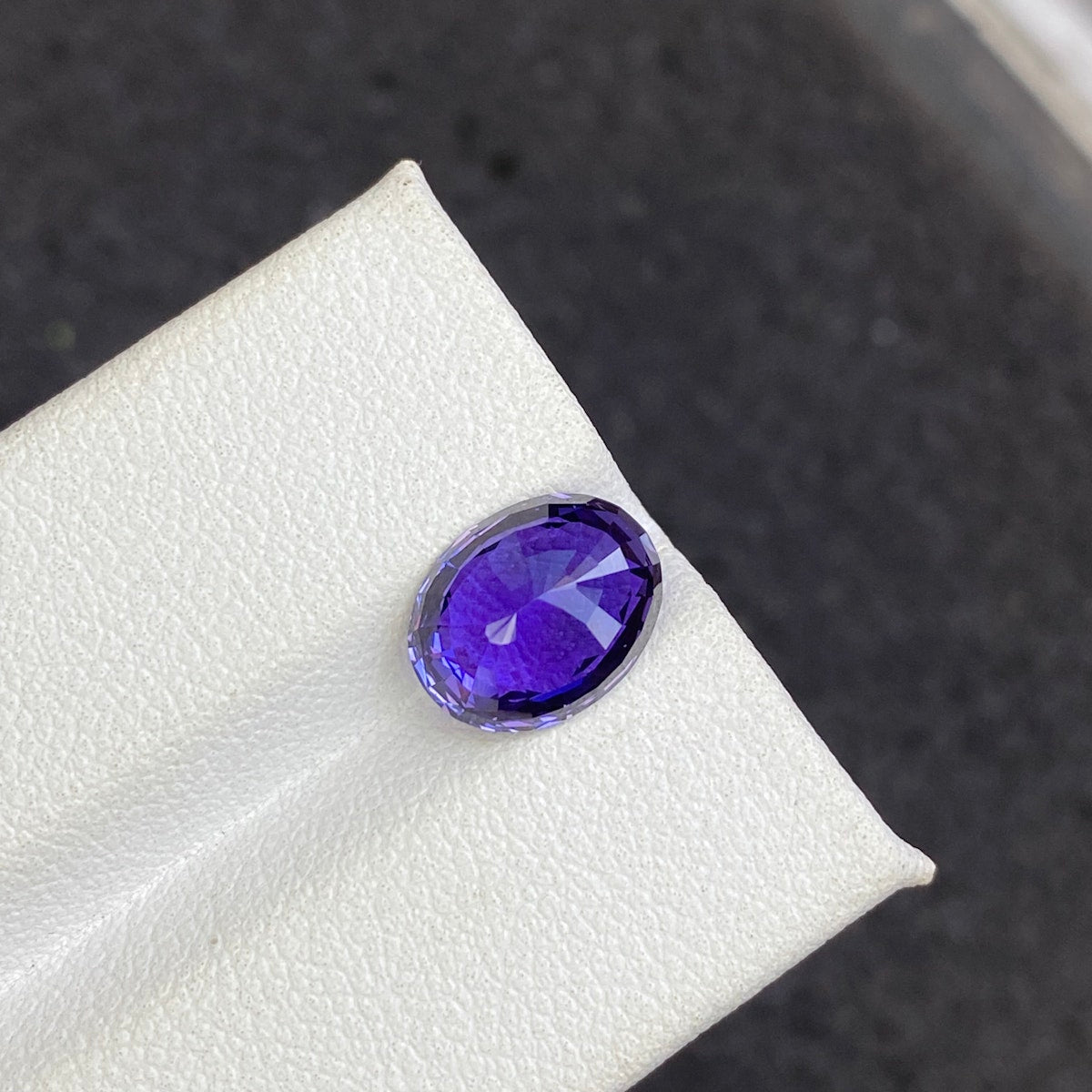 Un gros plan du Bonnot Paris Saphir Violet · 3,5 ct, une pierre précieuse violette facettée en forme de poire, repose sur une surface texturée blanche avec un pavé gris foncé flou en arrière-plan.