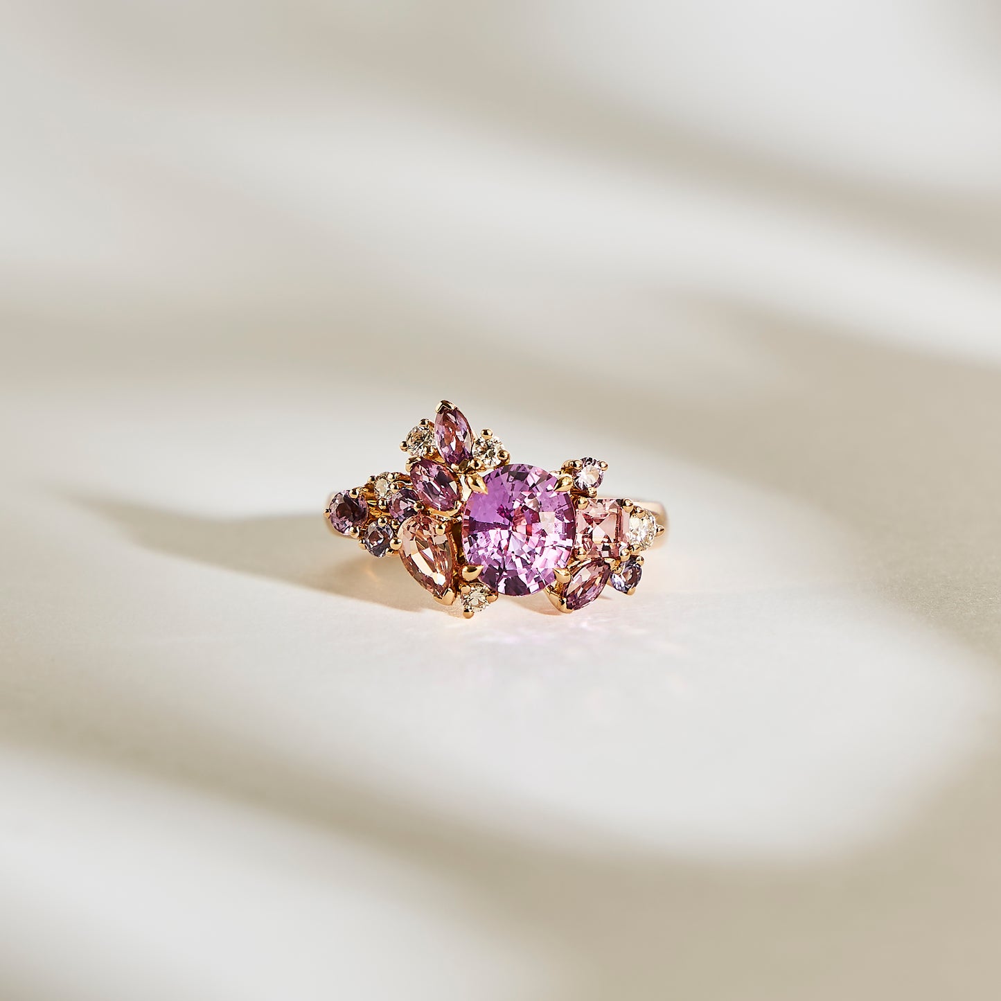 Le Purple Bloom - Saphir de Bonnot Paris présente une grande pierre précieuse rose entourée de diamants plus petits et de pierres claires, le tout serti sur une bande d'or, élégamment présentée sur une surface texturée doucement éclairée projetant des ombres douces.