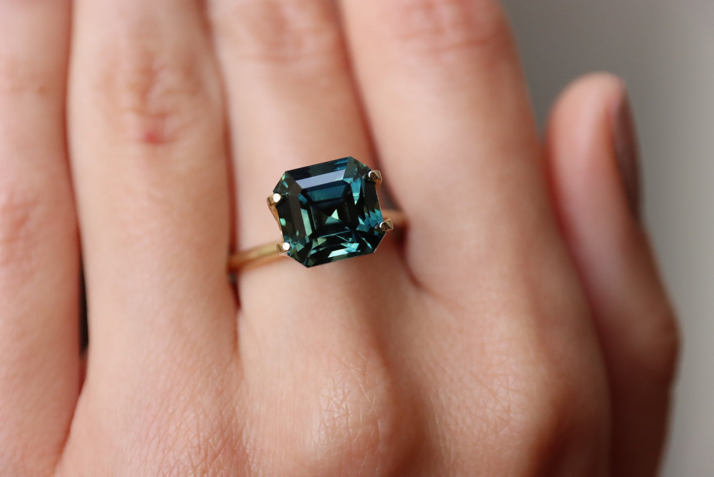 Un gros plan montre une main à la peau claire portant la bague en or Saphir Vert · 6,53 carats de Bonnot Paris avec une pierre précieuse verte taille émeraude sur le majeur, sur un fond légèrement flou.