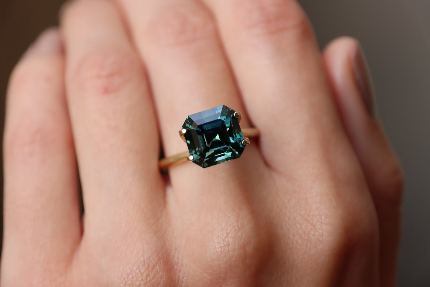 Une main met en valeur la bague en or Saphir Vert · 6,53 carats de Bonnot Paris, ornée d'une pierre turquoise taille émeraude sur un anneau lisse. Le flou de l'arrière-plan met en valeur les facettes détaillées de la bague.