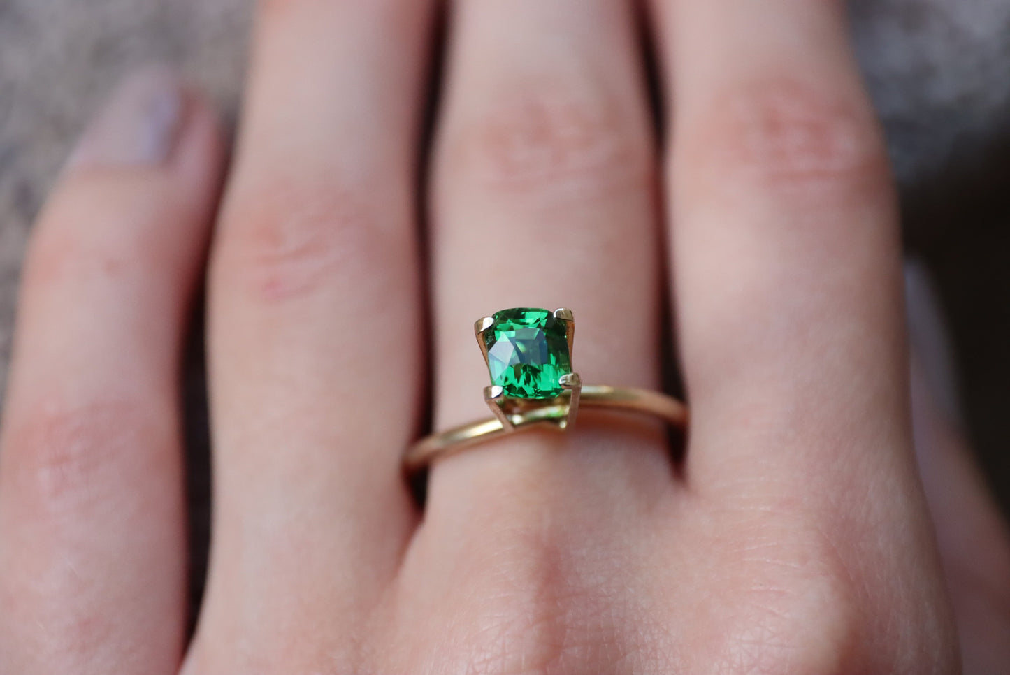 Une main ornée d'une bague en or Grenat Tsavorite · 1,55 carat de Bonnot Paris avec une pierre précieuse verte rectangulaire repose sur une surface douce et texturée.