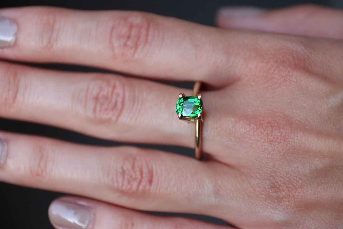 Une main ornée de Grenat Tsavorite Bonnot Paris, une pierre verte rectangulaire de 1,55 carat, se détache sur un fond sombre. Les ongles sont vernis d'une teinte claire et neutre.