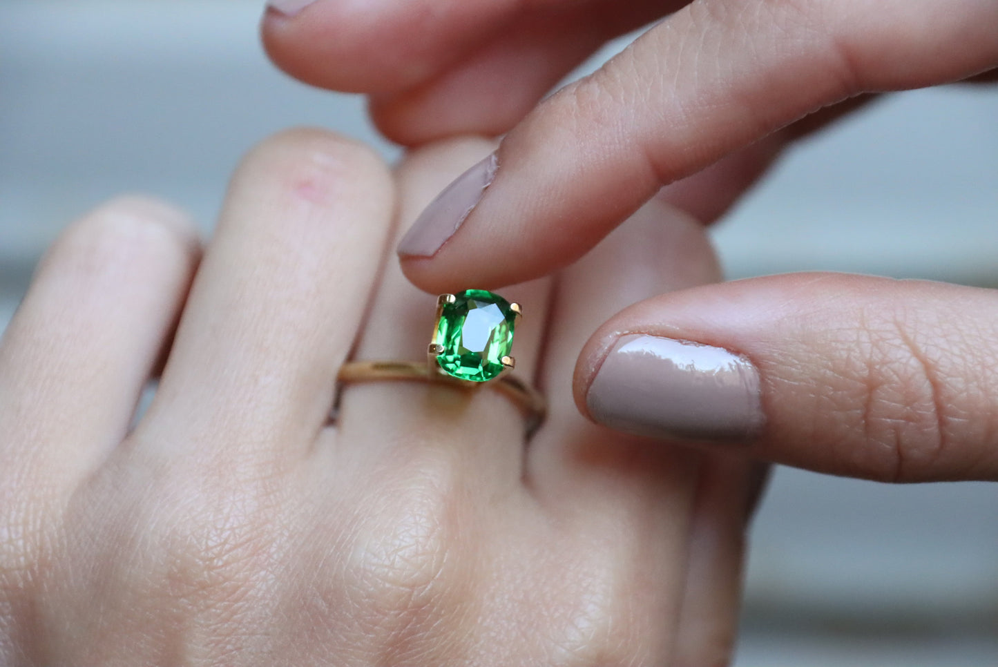 Un gros plan révèle une main portant un Grenat Tsavorite de Bonnot Paris, orné d'une pierre verte ovale de 1,7 carat. Une autre main, aux ongles parfaitement manucurés, effleure délicatement la pierre, soulignant sa couleur vive et sa clarté.