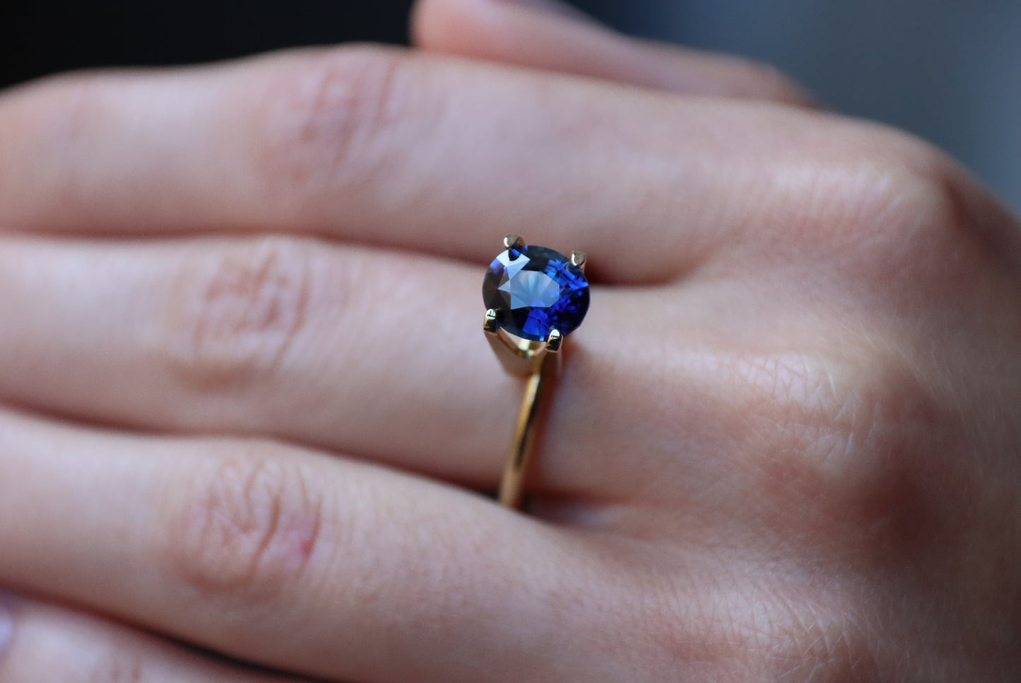 Une main porte la bague Saphir Bleu · 2,07 ct de Bonnot Paris, ornée d'une pierre précieuse ronde d'un bleu profond, reposant délicatement sur un fond légèrement flou.