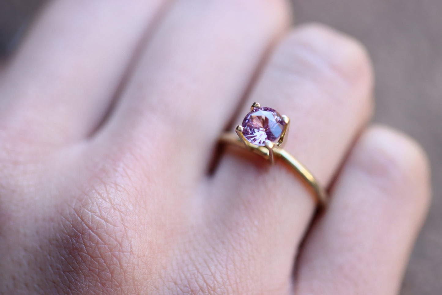 Un gros plan montre une main à la peau claire portant la bague Bonnot Paris Saphir Violet, ornée d'une pierre violet clair de 1,38 carat sertie à griffes. Son design élégant et simple reflète magnifiquement la lumière sur l'arrière-plan flou.