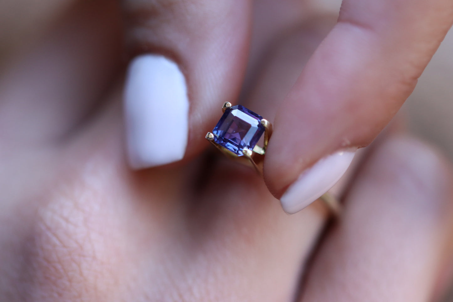 Une main, vernis à ongles blanc, tient délicatement une bague en or ornée de la pierre Saphir Violet de 1,65 carat de Bonnot Paris. L'accent est mis sur ses facettes et sa couleur intense.