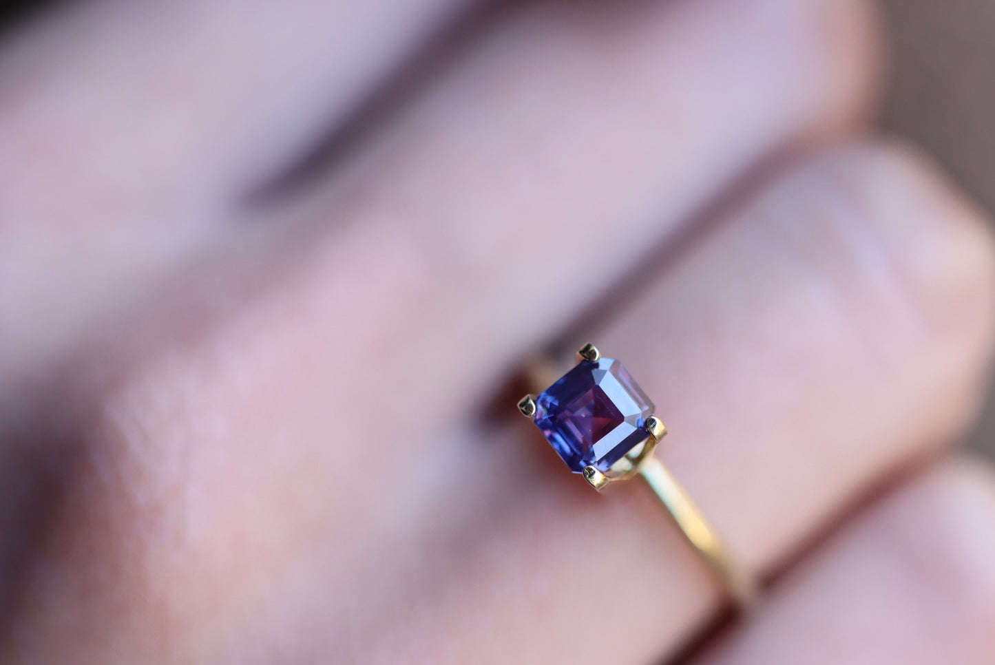 Gros plan d'une main portant une bague en or Saphir Violet Bonnot Paris avec une pierre précieuse taille carrée de 1,65 carat, sertie sur un fond légèrement flou qui met en valeur les superbes détails de la bague.