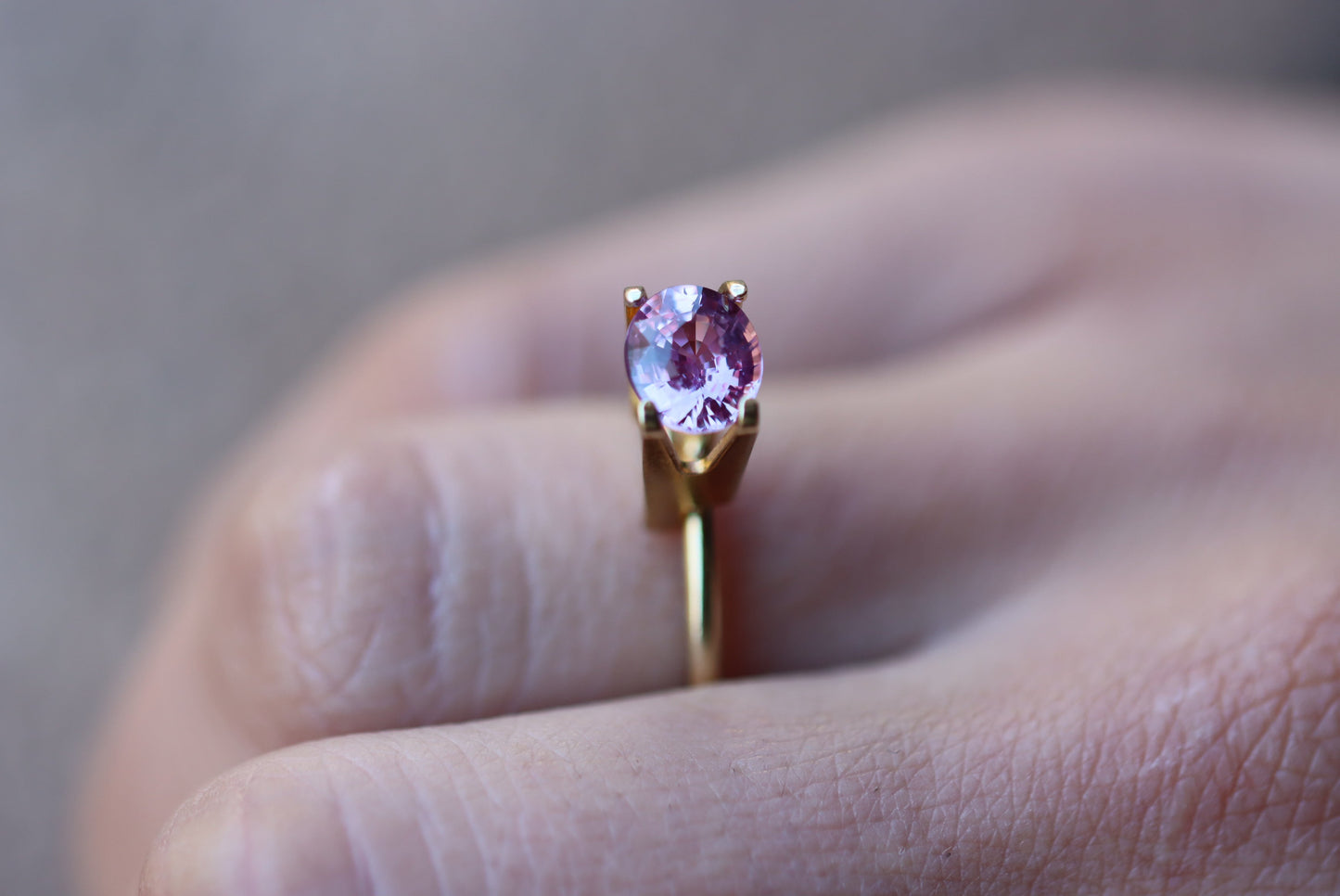 Une main ornée de la bague Saphir Violet · 1,61 carat en or de Bonnot Paris, mettant en valeur une pierre précieuse ronde et rose, est positionnée entre les doigts sur un fond légèrement flou.