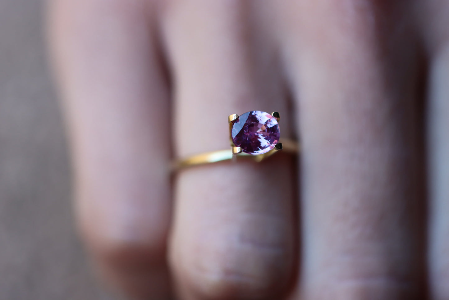 Un gros plan met en valeur une main portant la bague Saphir Violet en or 1,61 carat de Bonnot Paris, ornée d'une pierre violette à la taille scintillante, maintenue par quatre griffes. Elle se détache sur l'arrière-plan flou d'un doigt orné d'ongles neutres.