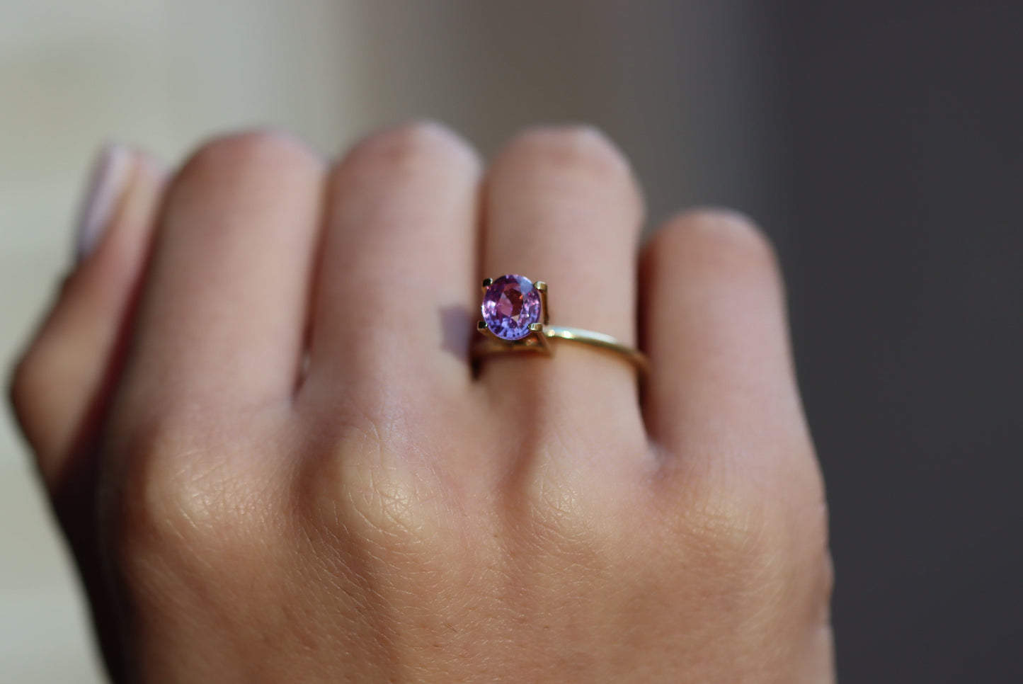 Une main ornée de la bague en or Saphir Violet · 1,51 carat de Bonnot Paris, ornée d'une pierre précieuse violette, repose sur le majeur, sur un fond légèrement flou.