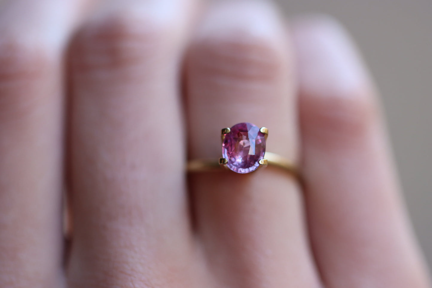 Une main ornée de la bague Saphir Violet · 1,58 carat de Bonnot Paris, ornée d'une pierre rose ovale, se détache sur un fond flou. La lumière tamisée accentue l'éclat de la pierre et l'élégance de son design.