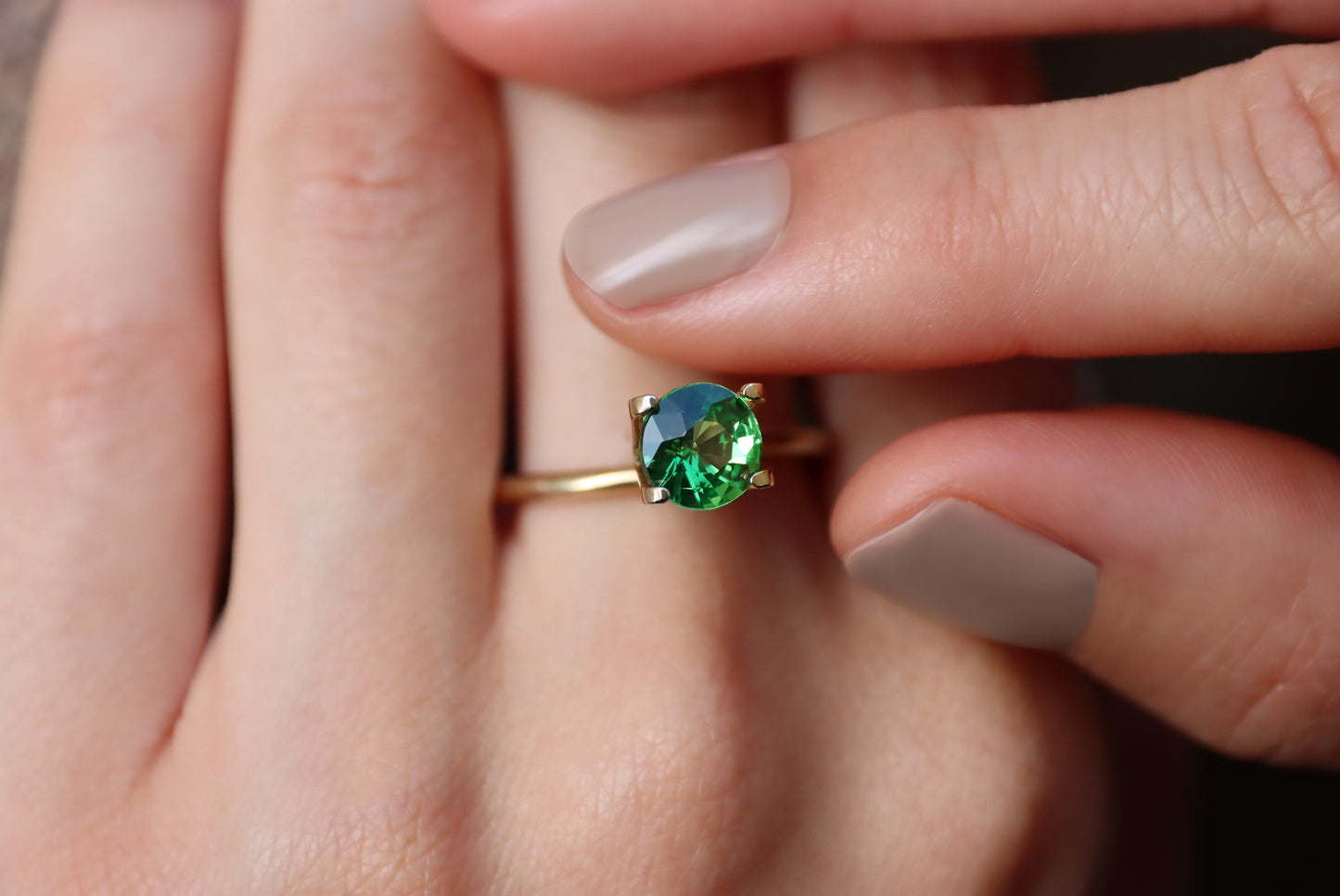 Des doigts soigneusement manucurés ajustent une bague en or ornée de la Grenat Tsavorite Bonnot Paris, une pierre précieuse verte ovale vibrante de 1,49 ct maintenue par quatre griffes, soulignant son design élégant et délicat.