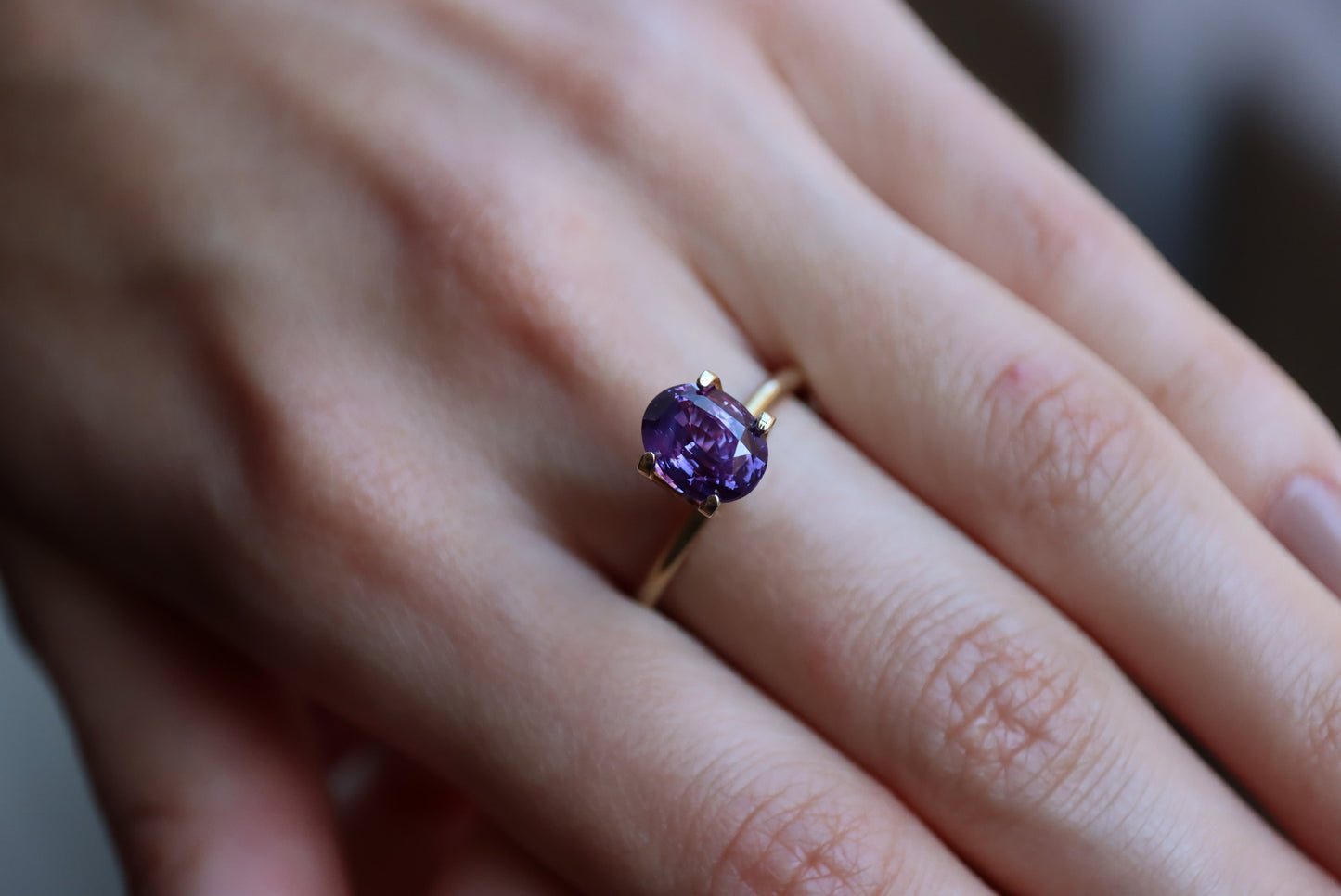 Gros plan d'une main à la peau claire portant la bague en or « Saphir Violet · 2,61 ct » de Bonnot Paris, ornée d'une pierre précieuse violette de forme ovale, sur l'annulaire sur un fond flou.